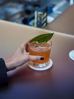 Hand holding an orange mocktail garnished with a bay leaf on a coaster at a bar in Le Pavillon.