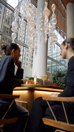 Two people seated inside a midtown bar with large glass windows and decorative hanging glass sculptures overhead.