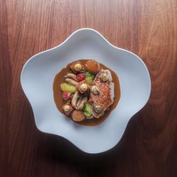Fine dining dish with fish, scallops, mussels, and garnishes in a uniquely shaped white plate on a wooden table.