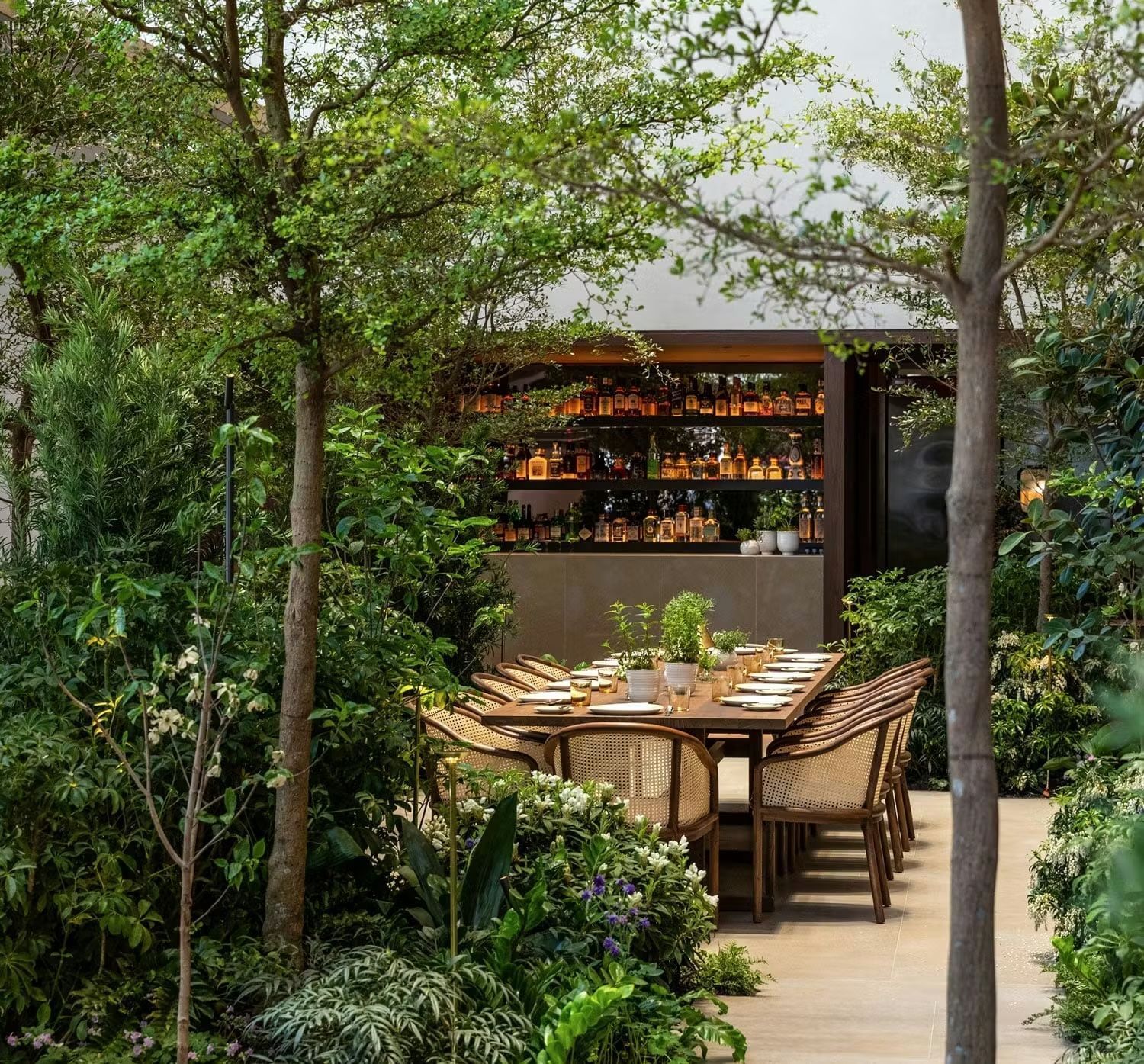 Aesthetic outdoor dining area featuring a wooden table surrounded by lush greenery and a well-stocked bar in the background.