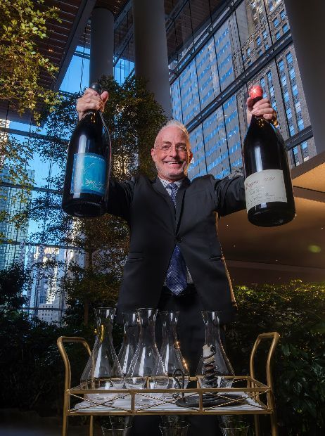 A waiter showcases two large bottles of wine in an elegant setting, with a cart of glasses nearby.