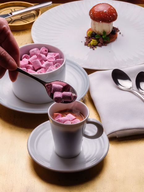 A cup filled with hot chocolate and topped with pink marshmallows, alongside a bowl of more marshmallows.